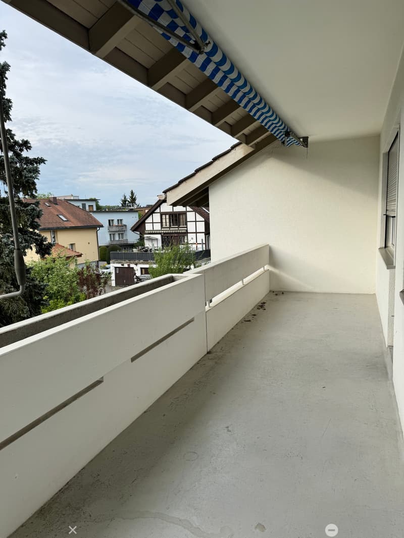 Balcony with white walls, concrete floor, and a blue and white striped awning extending from the roof.
