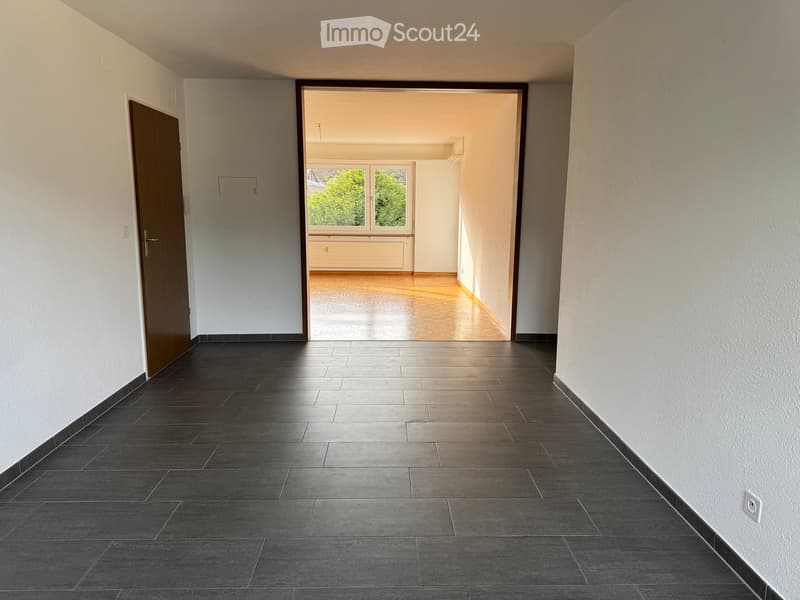Empty hallway with dark tiled floor, white walls, and a wooden door on the left. An open doorway leads to a room with wooden flooring and large windows showing greenery outside.