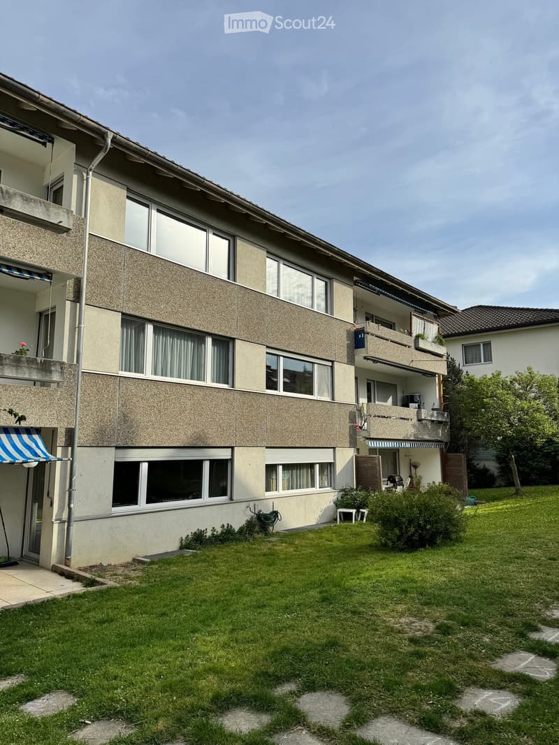Multi-story residential building with balconies, large windows, and a well-maintained lawn in front.