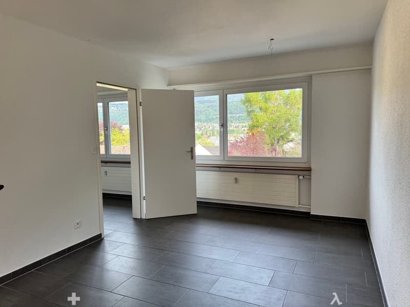 Empty room with dark tiled floor, white walls, large window with view, radiator, and open door leading to another room.