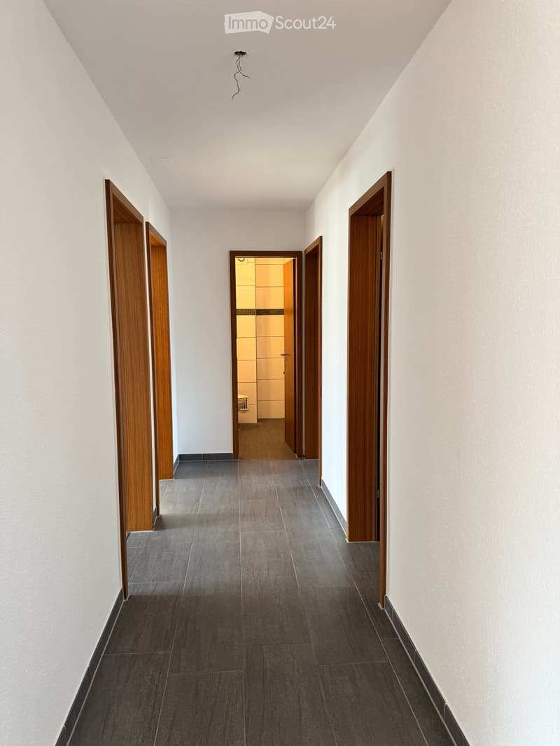 A clean, modern hallway with grey tiled flooring, white walls, and wooden doors on both sides. A small, open doorway at the end leads to a brightly lit room.