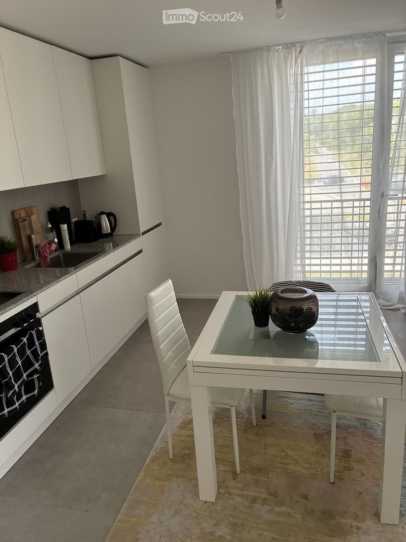 Modern kitchen with white cabinets, a glass dining table with a white chair, and various appliances including an oven, sink, and coffee maker. There is a vase with a plant on the table and a curtain covering the window.