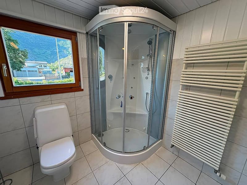 Modern bathroom with a corner shower, toilet, and a white radiator. The shower has multiple spray heads and a glass enclosure. A large window offers a view of the outdoors.