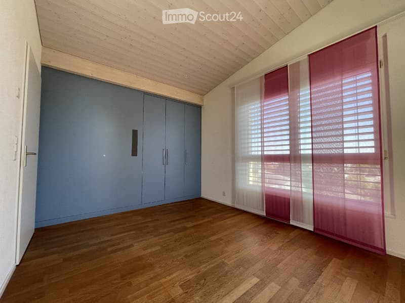 Empty room with wooden flooring, white walls, and a large built-in wardrobe with grey doors. Windows with red blinds are visible on one side.