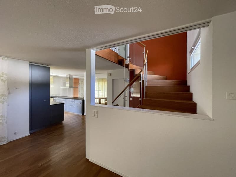 Modern hallway with wooden flooring, glass staircase railing, and visible kitchen area in the background.