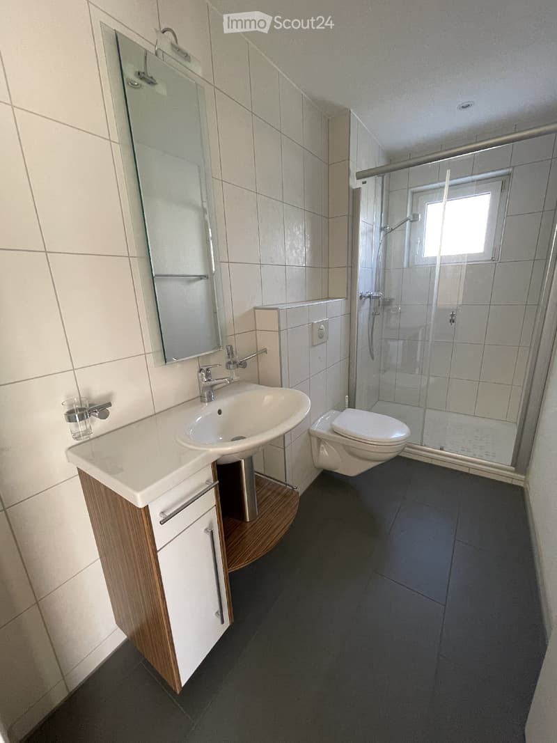 Modern bathroom with a white sink, toilet, shower, and mirror. The sink has a wooden cabinet underneath, and a small glass cup is placed on the sink counter.