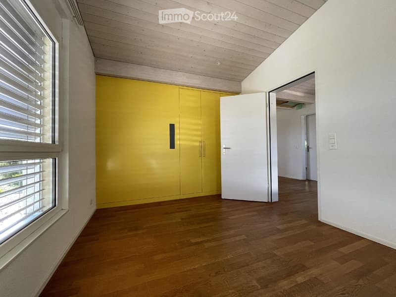 Empty room with wooden flooring, white walls, and a large window with blinds. A yellow cabinet is visible, and an open door leads to another room.