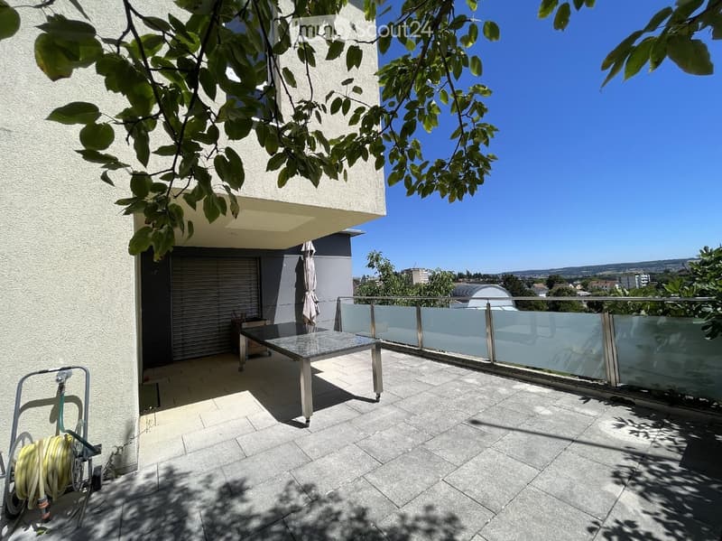 Balcony with tiled floor, glass railing, and a small table. The balcony is attached to a modern building with a view of the surrounding area.