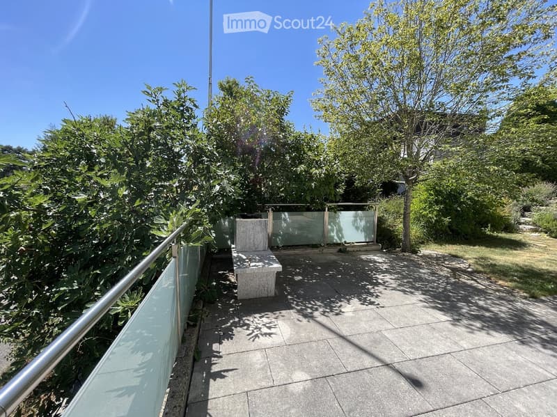 Balcony with a modern glass railing, stone bench, and lush greenery in the background.