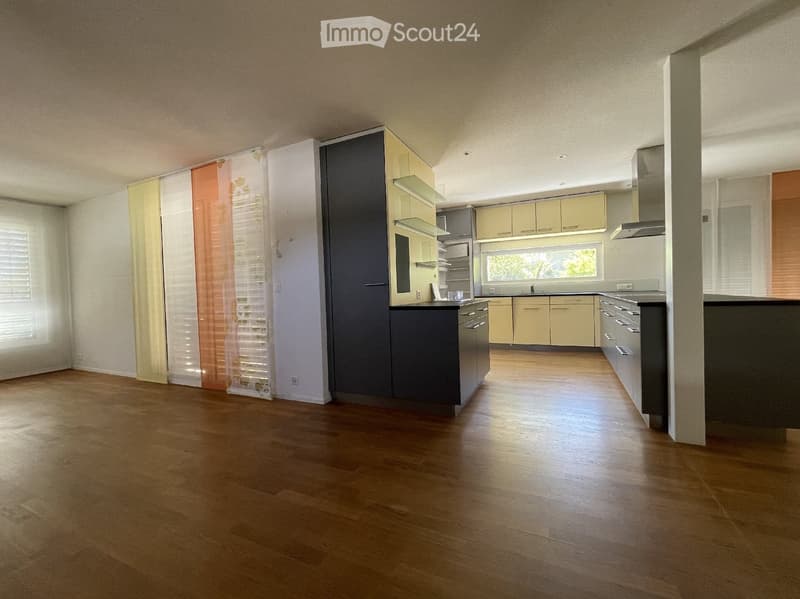 Empty kitchen with light-colored cabinets, dark countertops, and wooden flooring. The space is well-lit with large windows and modern fixtures.