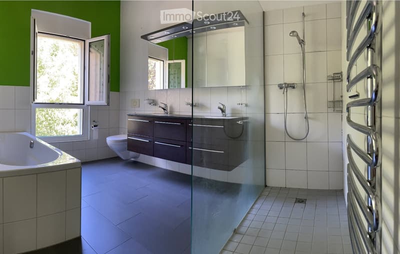 Modern bathroom with a bathtub, toilet, double sink, large mirror, and a glass-enclosed shower area. The space features tiled walls and floors, a radiator, and a window with green accents.
