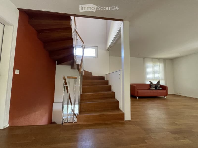 A modern living room with wooden flooring, a staircase with wooden steps and metal railings, and a red sofa with decorative pillows.