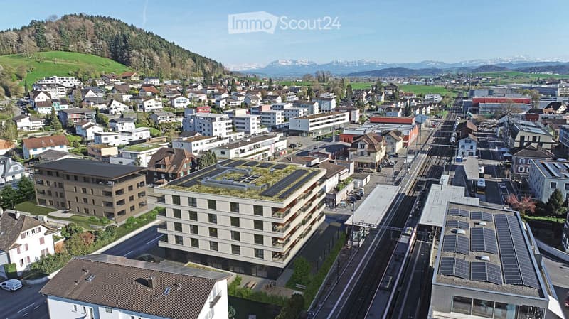 Aerial view of a town with multiple buildings, including residential houses, commercial structures, and a modern building with a green roof and solar panels. The town is surrounded by green hills and mountains.