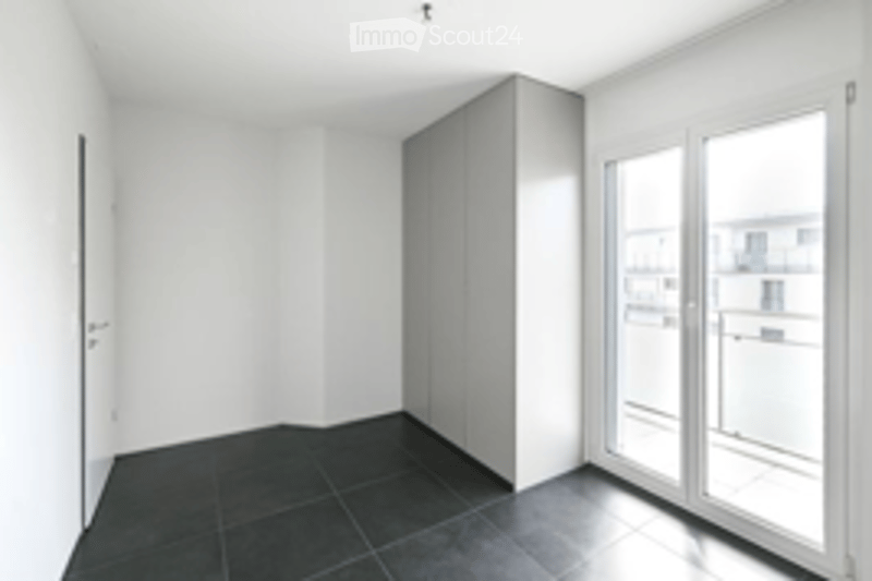 Empty room with white walls, dark tiled floor, large glass sliding doors leading to a balcony, and a built-in wardrobe unit.