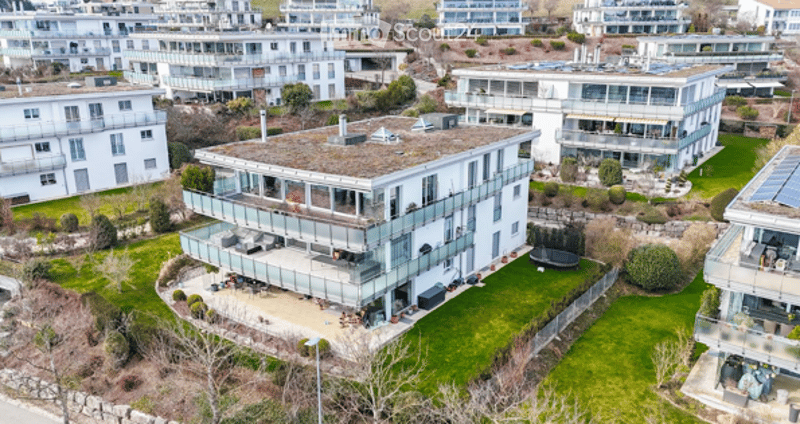 Aerial view of a modern multi-story residential building with a flat roof, large windows, and a spacious balcony area. The building is surrounded by well-maintained lawns and neighboring houses.