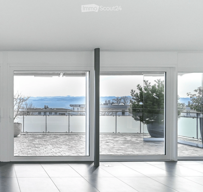 Bright, minimalist room with large glass doors leading to a balcony, offering a scenic view of the city and mountains. The space features a clean, white interior with a tiled floor and modern design.