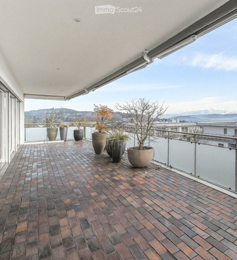 Modern balcony with brick tile flooring, glass railing, and several potted plants arranged along the edge, offering a scenic mountain view.