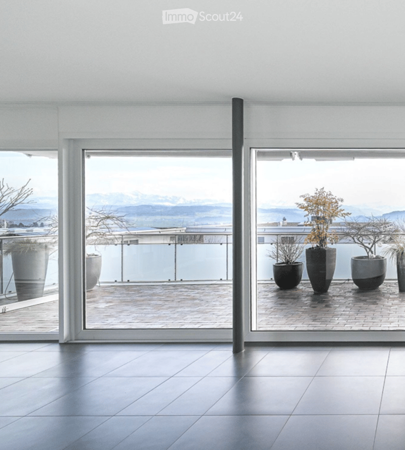 Modern balcony with large glass doors, tiled floor, and a scenic mountain view. Potted plants are placed on the balcony outside the doors.