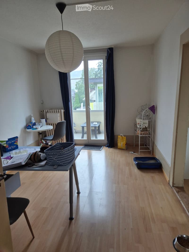 A furnished room with wooden flooring, a large hanging lamp, a desk with a chair, a small table with a chair, a shelf, and a cat litter box.