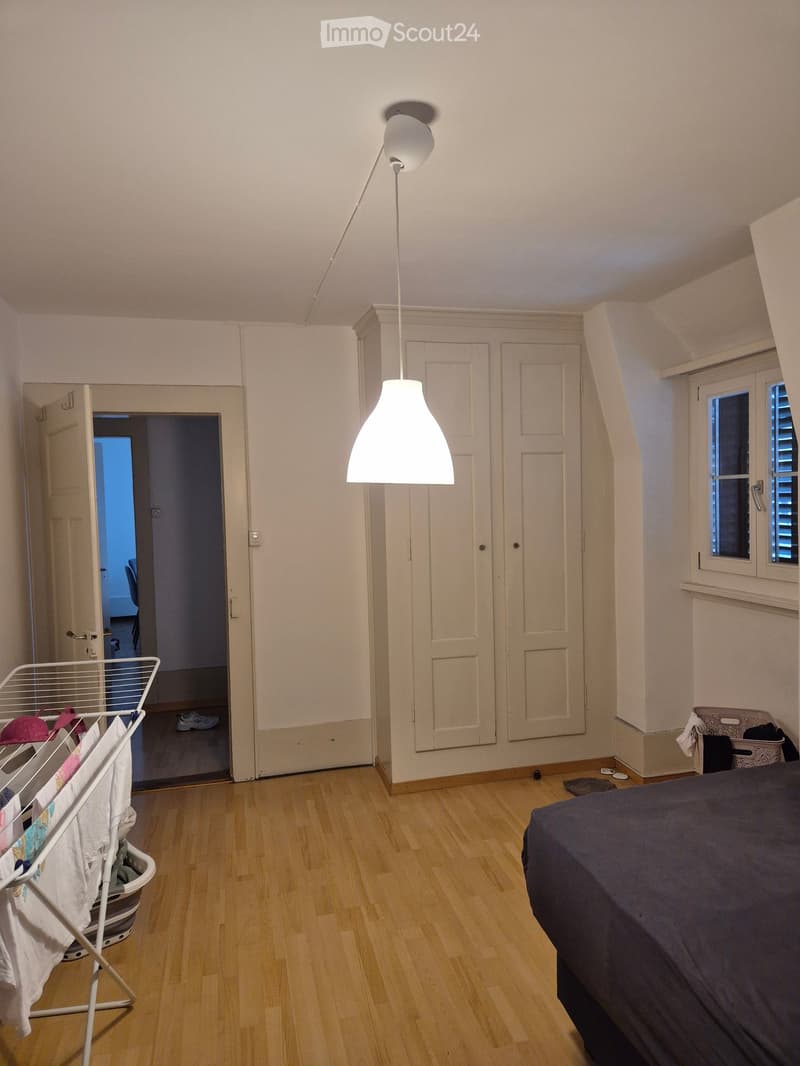 Bright bedroom with wooden flooring, a large bed with dark bedding, a wardrobe, a hanging lamp, a laundry basket, and a chair visible in the adjacent room.