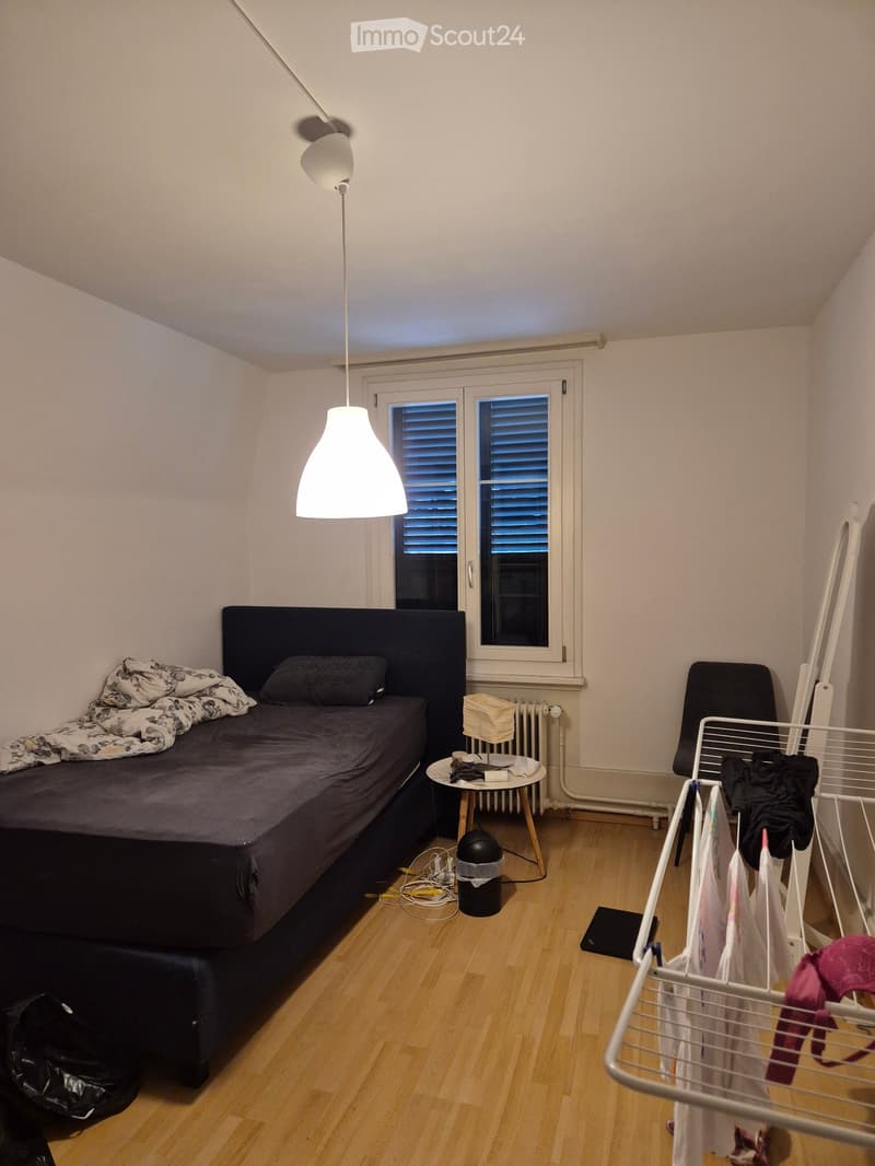 A small, furnished bedroom with a bed, nightstand, chair, and a laundry basket. The room features wooden flooring and a window with blinds.
