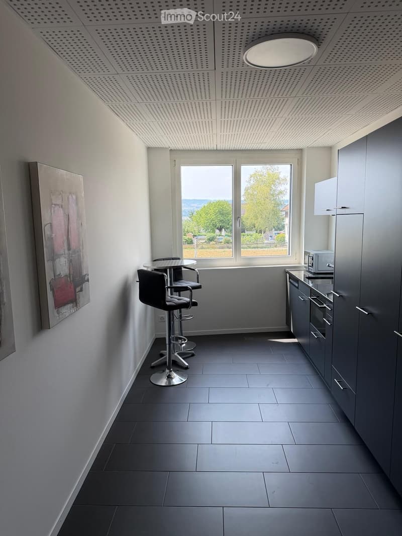 Modern kitchen with grey cabinets, a microwave, a small round table with four stool chairs, and a large window offering a view of greenery outside.