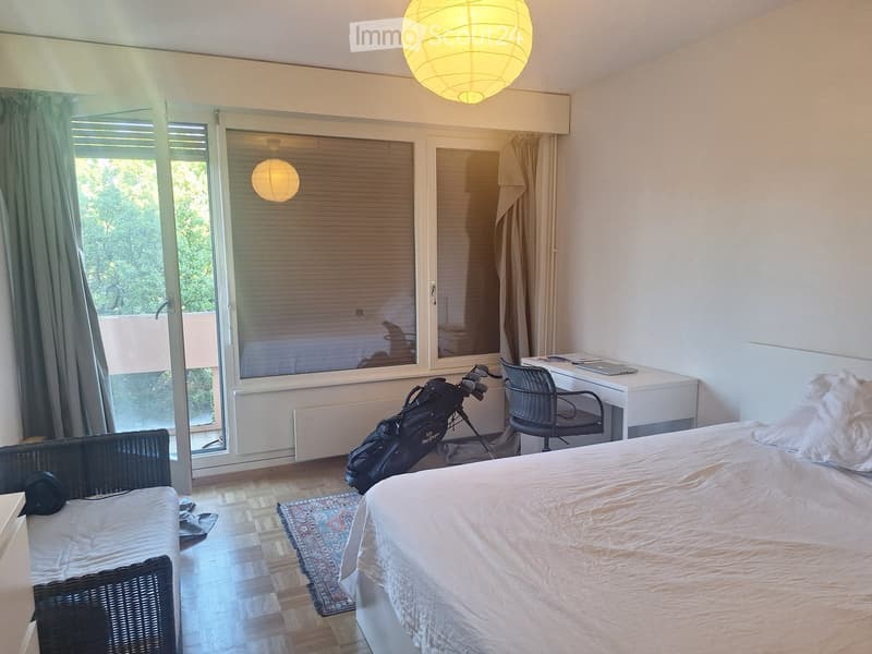 Bright bedroom with a large bed, wicker sofa, desk, and golf bag near the window. The room features wooden flooring, a patterned rug, and large windows with blinds.