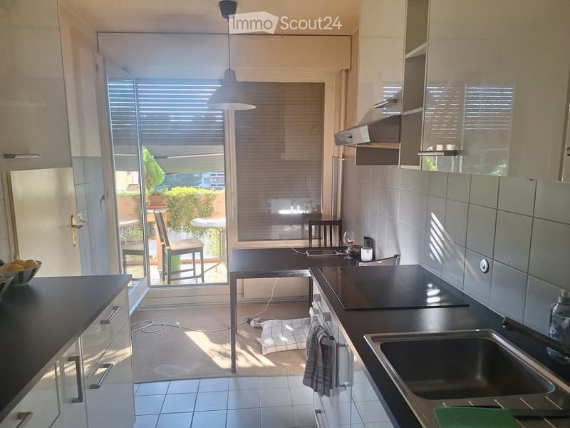 Modern kitchen with dark countertops, stainless steel sink, and built-in cabinets. A small dining area with a table and chairs is visible through a large window with blinds.