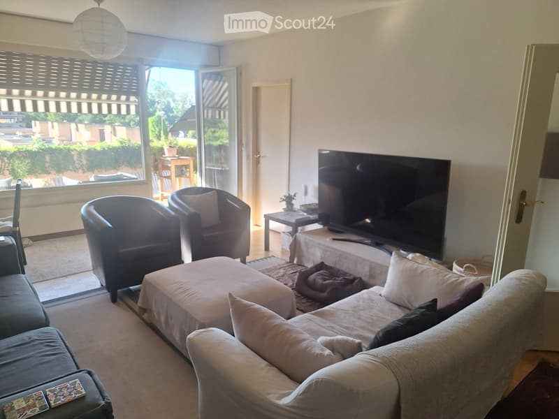 Bright living room with large sliding glass doors leading to a balcony, a grey sectional sofa with an ottoman, two armchairs, a coffee table, a television on a stand, and a side table with a lamp.