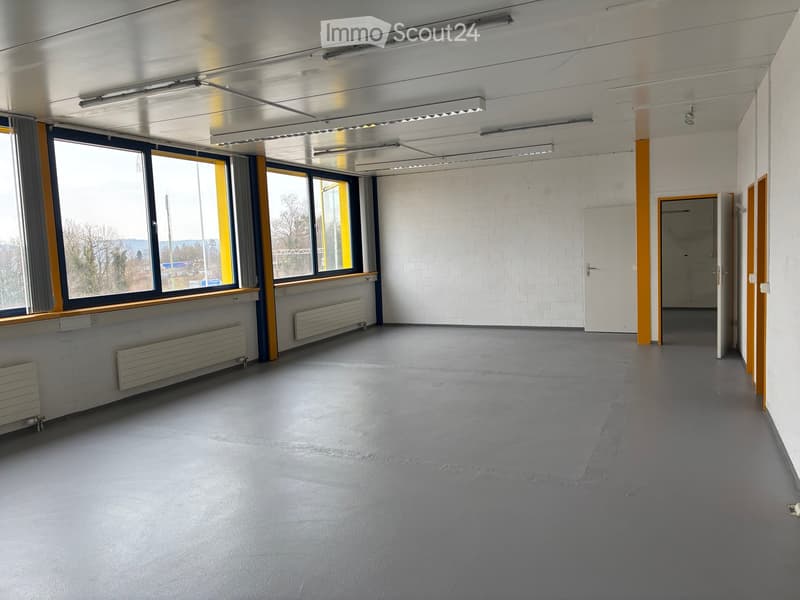 Empty commercial space with concrete flooring, large windows, heaters, and open doorways.