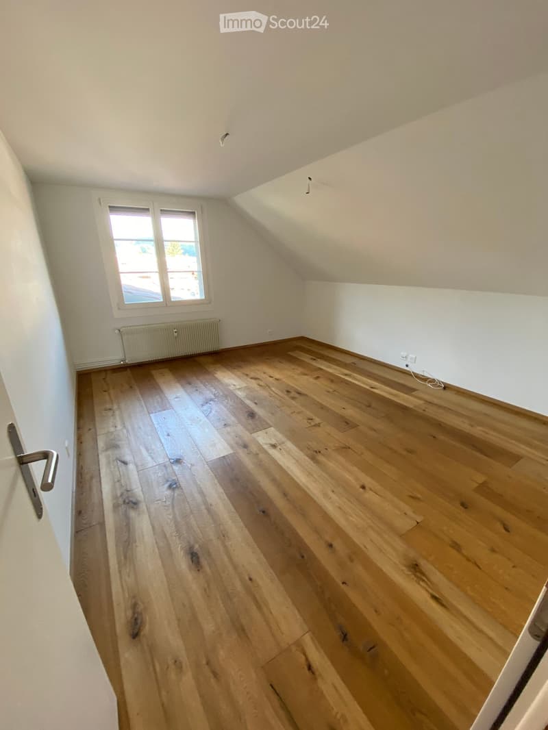 Empty room with wooden flooring, white walls, and a window with a radiator below it. The room has a slanted ceiling typical of an attic space.