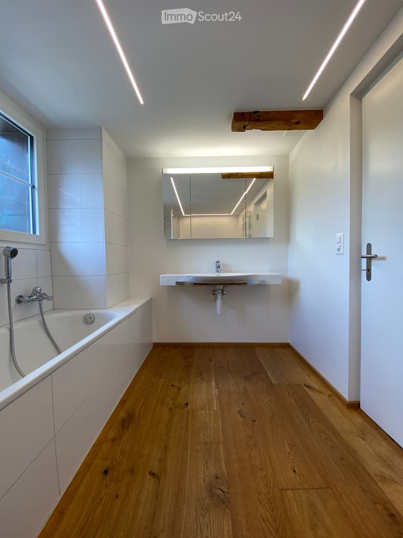 Modern bathroom with wooden flooring, white tiled walls, a bathtub, a sink with a mirror, and a window providing natural light.