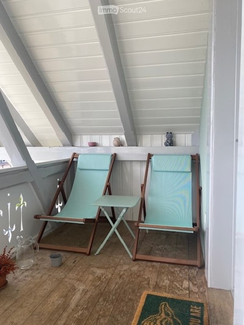 A wooden balcony with a slanted white ceiling, two light blue folding chairs, a small table, and decorative items on the railing.