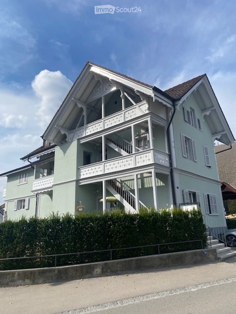 Two-story house with light green exterior, white trim, and multiple balconies. The house features decorative railings and a well-maintained hedge in front.