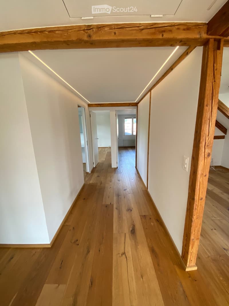 A long, narrow hallway with wooden flooring and white walls, featuring exposed wooden beams and recessed lighting. The hallway leads to several open doorways, with visible wooden stairs on the right side.