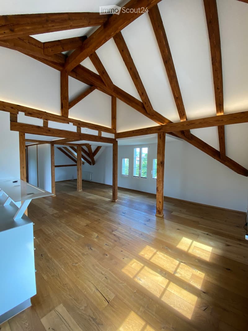 Bright, open attic space with exposed wooden beams and wooden flooring. Large windows allow natural light to fill the room. The space is completely empty, with no furniture or furnishings visible.
