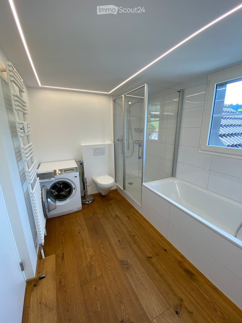 Modern bathroom with wooden flooring, white tiled walls, a bathtub, a shower, a toilet, and a washing machine. Features a large window and built-in radiator.