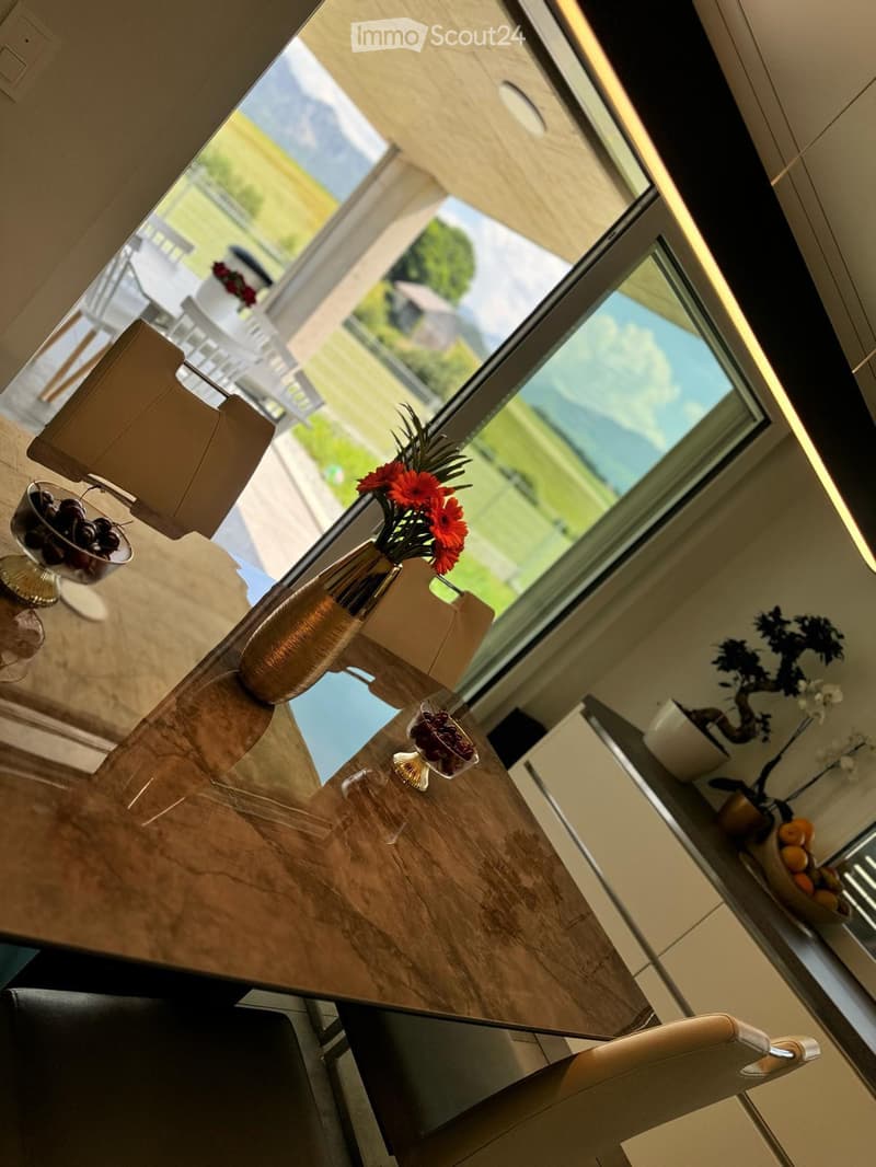Modern kitchen with a wooden table, chairs, vase with flowers, and fruit bowls, overlooking a scenic outdoor view.