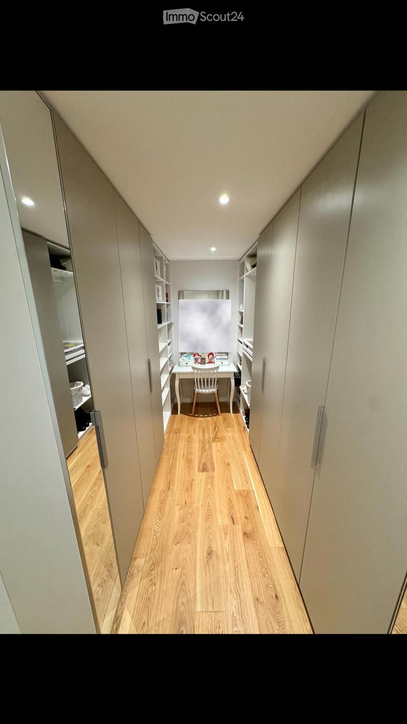 A narrow hallway with wooden flooring leads to a small, furnished room containing a desk, chair, and mirror. The room features built-in shelves and a sleek, modern design.
