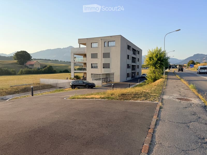 Modern multi-story building with balconies and large windows, surrounded by a parking area and open landscape with distant mountains.