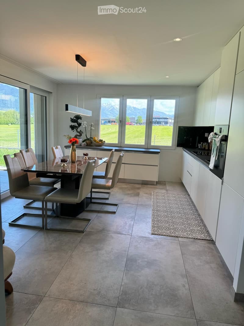 Modern kitchen and dining area with white cabinets, a glass dining table, six chairs, a vase with flowers, and a bowl of fruit on the counter. Large windows offer a view of a green field and mountains.