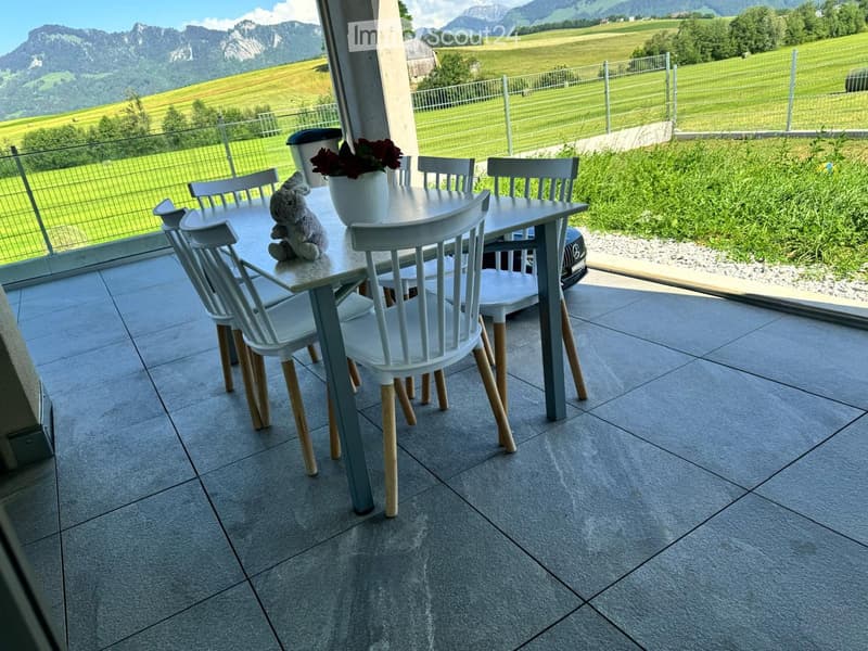Balcony with a dining table, chairs, vase with flowers, and a stuffed toy, overlooking a scenic mountain and green field view.