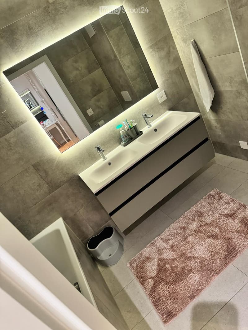 Modern bathroom with a large sink, mirror, and a pink bath mat. A small stool is placed near the bathtub, and a toothbrush holder sits on the sink counter.