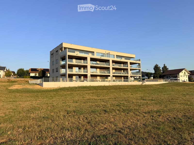 Modern multi-story residential building with balconies and large windows, surrounded by a grassy field.