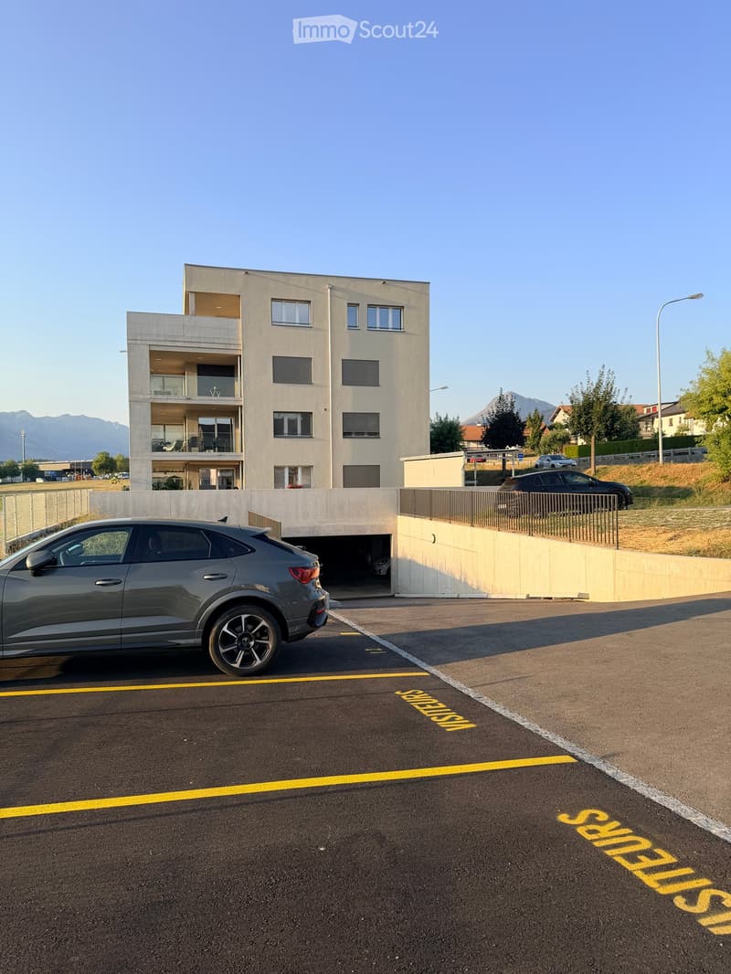 Modern multi-story building with balconies and large windows, surrounded by a parking area with several cars and a clear view of mountains in the background.