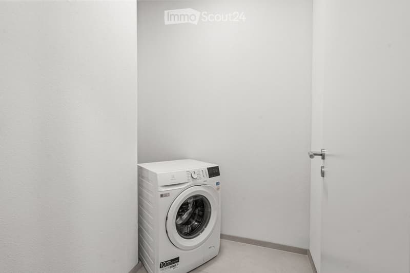 A small, empty room with white walls and a tiled floor, featuring a single white washing machine placed against the wall.