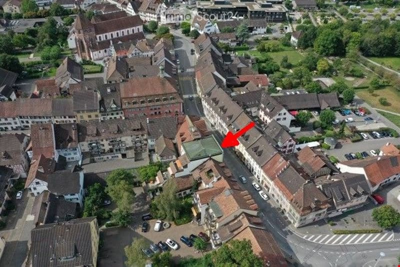Aerial view of a town with many buildings, some with brown roofs, a road with cars parked on both sides, and a red arrow pointing to a specific building.