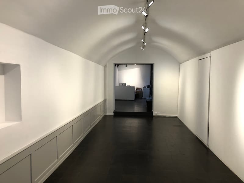 A long, narrow hallway with white walls, dark tiled flooring, and recessed lighting. The hallway leads to an open area with a reception desk and office furniture visible in the background.