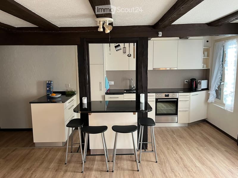 Modern kitchen with white cabinets, black countertop, bar stools, and stainless steel appliances including an oven and a faucet. Wooden floor and exposed wooden beams on the ceiling.