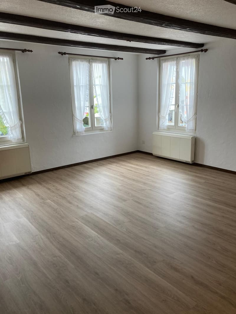 Empty room with wooden flooring, white walls, and large windows with white curtains. Radiators are visible beneath the windows.